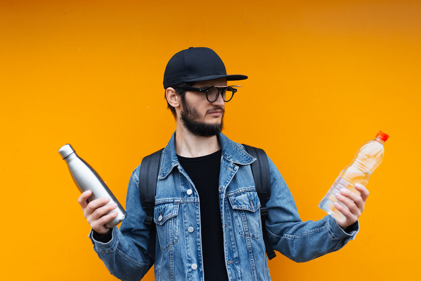 Portrait of cheerful young hipster, holding steel thermo eco bottle for water and plastic bottle, on yellow or orange background. Say No to Plastic.